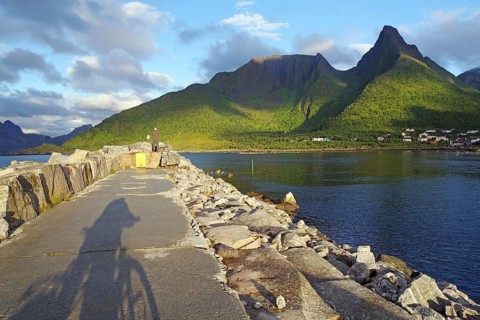 A stone path along the sea with a bicycle shade. The view leads to green mountains, partly sunny