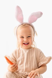 A young girl wearing bunny ears smiles joyfully while holding a carrot. She participates in Easter