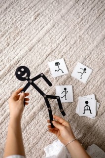 Hands positioned with a stick figure toy and several cards lying on a carpeted floor