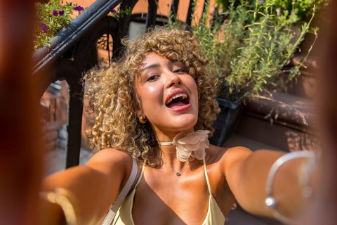 Joyful woman with curly hair captures a selfie on a brownstone stoop, enjoying urban summer life in