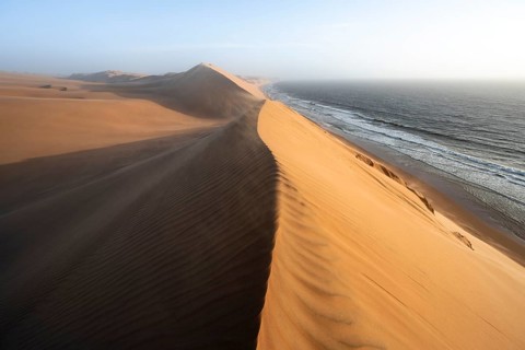 Sand dunes with wave patterns and sea in evening light, wind blowing sand across the dunes,