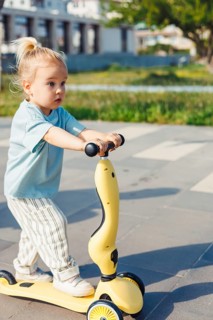 A young child with a ponytail rides a yellow scooter on a paved path in a park. Surrounding