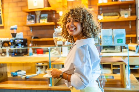 Smiling woman with curly blonde hair holding a cup of coffee inside an urban cafe, reflecting a