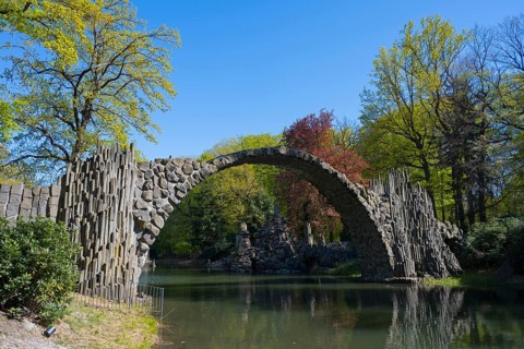Stone bridge against colorful, wooded background on a sunny day, over a calm body of water, Rakotz