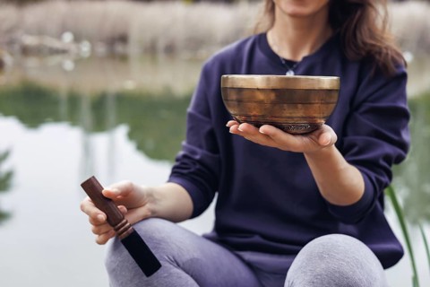 A young Caucasian woman with long brown hair holds a wooden bowl near a serene lake. Trees and