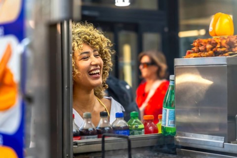 Young woman with curly hair laughing joyfully behind a food cart, offering a glimpse into the