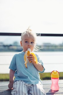 Young child toddler sits on a bench by the water, happily eating healthy snack banana. Sun shines
