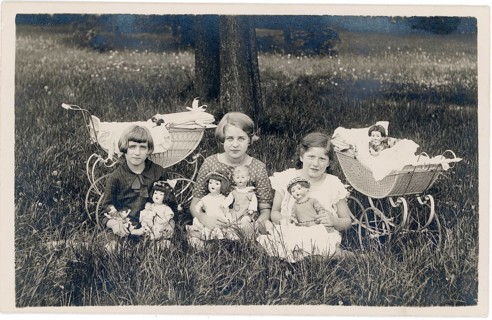 Three girls sitting with dolls and doll prams in an outdoor meadow, well-dressed, middle-class,