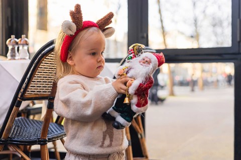 A young Caucasian girl with blonde hair wears a reindeer headband. She holds a Santa Claus toy in a