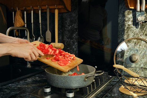 Pouring tomatoes from wooden board into pan. Pan is on stove filled with simmering vegetables