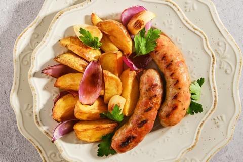 Two grilled sausages sit beside roasted potatoes and pieces of onion on decorative plate. Fresh