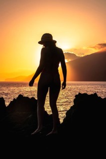 Woman wearing a straw hat and bikini standing on volcanic rocks, silhouetted against a golden