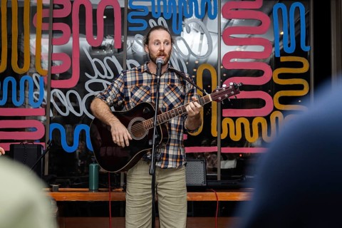 A young Caucasian man with a beard plays an acoustic guitar while singing on stage. Colorful