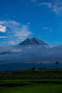 A majestic volcano rises above a green landscape surrounded by clouds under a clear blue sky,