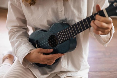 Person in white shirt playing a ukulele while seated on a wooden floor