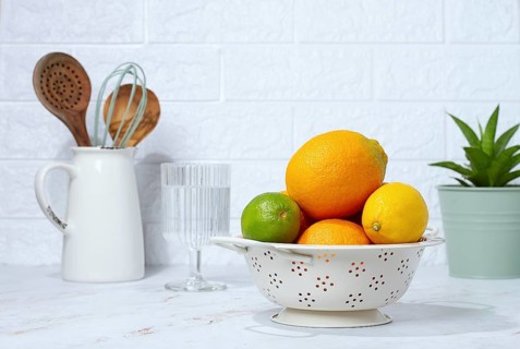 A bowl holds different fruits including oranges, lemons, and a lime on a table. Kitchen tools are