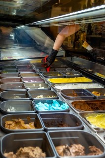 Hand wearing black glove preparing to scoop colorful gelato from a display case, showing a variety
