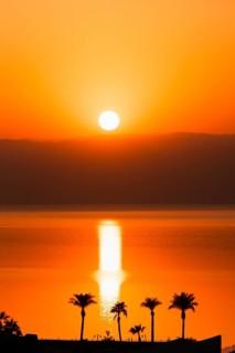 Dead Sea Jordan sunset with orange glow, calm water reflection, palm silhouettes, peaceful travel