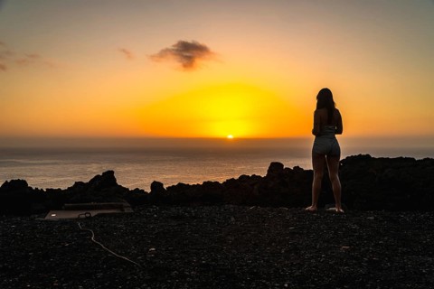 Woman standing on the volcanic rocky terrain watching a vibrant orange sunset over the atlantic