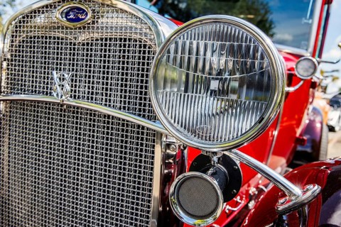Chrome grille, oval badge and drum headlamp of a vintage red Ford at a car meet