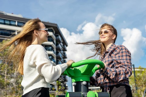 Two young women engage in outdoor exercise at a fitness station in an urban park. One has long