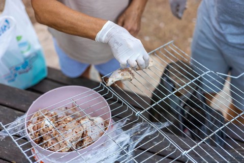 A person with brown skin and gloves prepares food on a grill. A bowl of marinated meat is nearby.