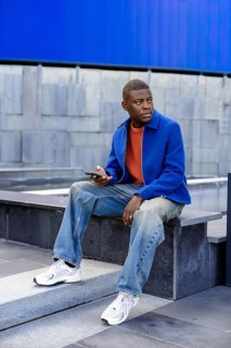 A young Black man sits on concrete steps, wearing a blue jacket and orange shirt. He holds a