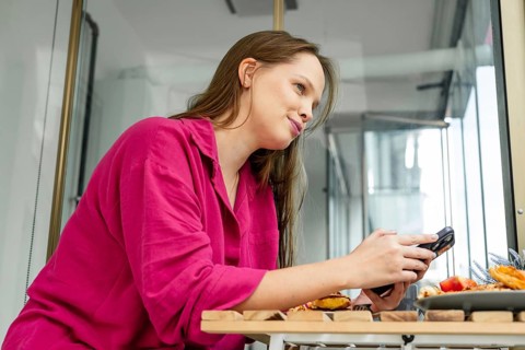 A young Caucasian woman with long brown hair sits at a table, holding a smartphone. She wears a