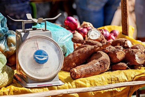 Market stand with cassava and onions on yellow cloth, next to a large scale, market scene in a