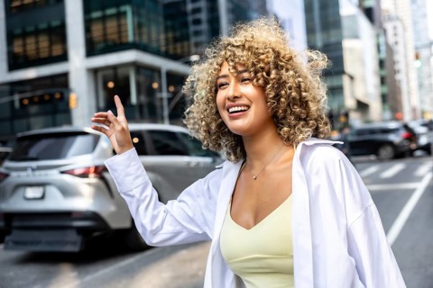 Young woman with curly hair smiling and raising her hand to hail a taxi, embodying urban lifestyle