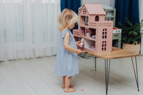 Child in a blue dress standing in a bright room playing with a dollhouse on a wooden table