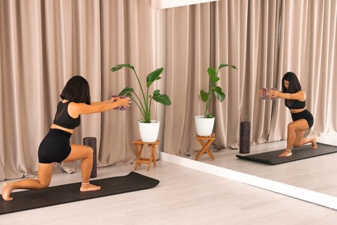 A woman doing lunges with dumbbells, mirrored in front of her, on a yoga mat