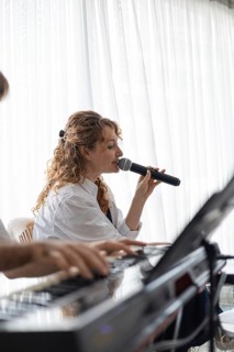 A young Caucasian woman with curly hair sings into a microphone. A hand plays a keyboard in the