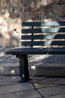 A dark green wooden bench stands beside a rough stone wall and steps in dappled light,