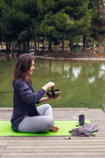 A young Caucasian woman with long brown hair sits cross-legged on a green yoga mat by a lake. She