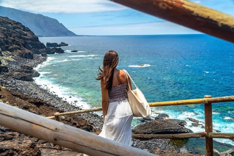 Woman standing on volcanic coastline watching tropical ocean waves and distant mountains,