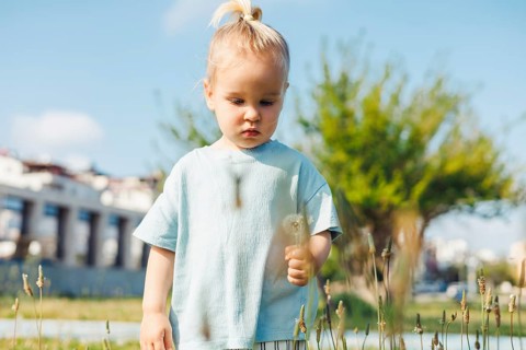 Young child in a light blue shirt crouches in a lush green field, intently picking dandelions. The