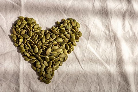 Green pumpkin seeds arranged in a heart shape on a white fabric background. The scene conveys love