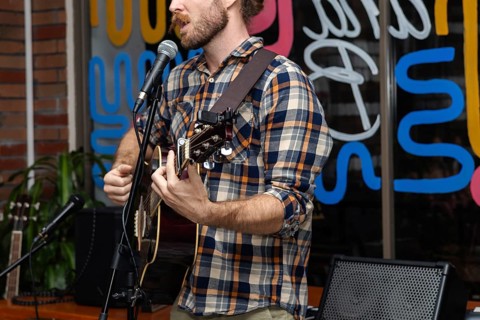 A young Caucasian man with a beard sings and plays an acoustic guitar in a vibrant indoor setting.