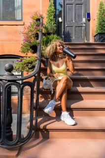 Young woman with curly hair sitting on brownstone steps, enjoying a refreshing drink from a