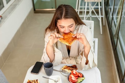 A young Caucasian woman with long brown hair enjoys a pastry at a table. A cup and a plate with