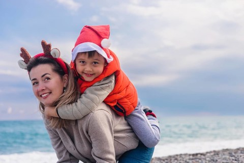 A young Caucasian woman with brown hair wears reindeer antlers. A young Asian boy in a Santa hat