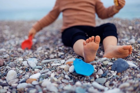 Two years girl toddler sits on a pebbly beach, playing with a yellow bucket and blue shovel.