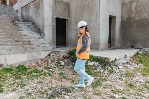 A young Hispanic woman in a safety vest and hard hat walks on a construction site. The background