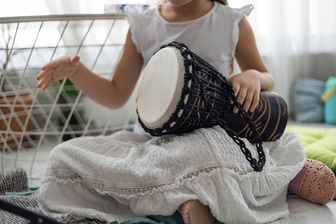 A young girl plays a black drum at home. She wears a white dress and sits on a colorful mat. The