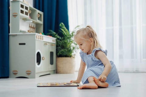 Child in a blue dress sitting on the floor playing with a puzzle in a bright playroom with a toy