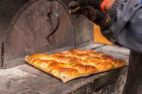 A baker removes a tray of freshly baked golden pastries from a traditional oven. The pastries have