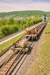 Workers wearing orange clothing working on tracks in a tree-lined hillscape, Leonhard Weiss