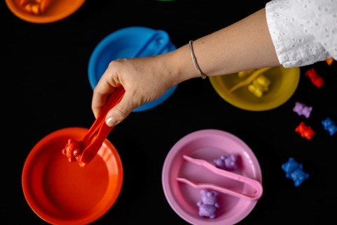 A hand sorts bears using tongs into colorful plates on a dark surface