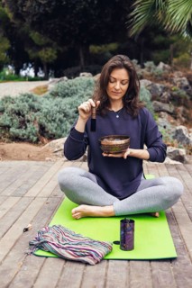 A young Caucasian woman with brown hair plays a singing bowl outdoors. She wears a dark sweater and